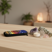 A black zippered pouch with a colorful cosmic and temple design lies on a light wood table next to a lit candle, crystals, and a smudge bundle, with a softly blurred home decor background.