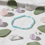A light blue beaded bracelet arranged in a circle on a white marble surface, surrounded by green leaves, pink and purple crystals, and clear crystal points.