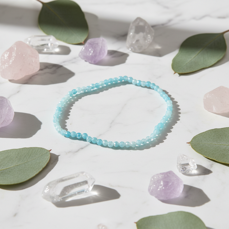 A light blue beaded bracelet arranged in a circle on a white marble surface, surrounded by green leaves, pink and purple crystals, and clear crystal points.