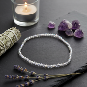 A beaded bracelet made of clear faceted beads arranged in a circle on a dark slate surface, styled with lavender sprigs, raw amethyst crystals, a smudge bundle, and a lit candle in a glass holder.