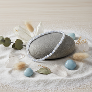 A beaded bracelet made of pale blue-white round beads draped over a smooth gray stone, styled on white sand with crystals, dried grass, and small blue stones.
