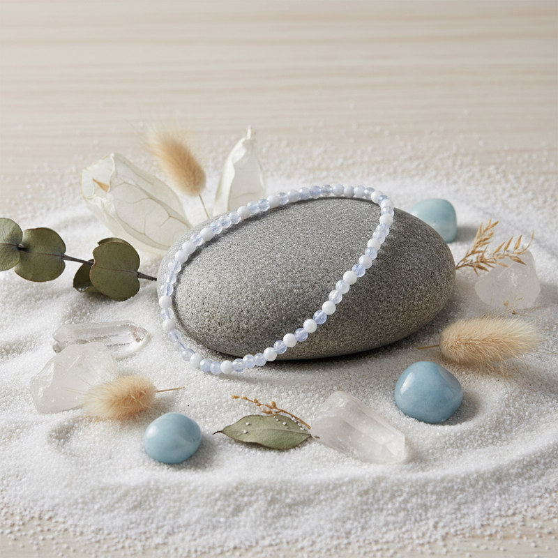A beaded bracelet made of pale blue-white round beads draped over a smooth gray stone, styled on white sand with crystals, dried grass, and small blue stones.