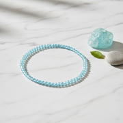 A light blue beaded bracelet arranged in a circle on a white marble surface, with a matching translucent blue stone and a smooth white stone nearby.