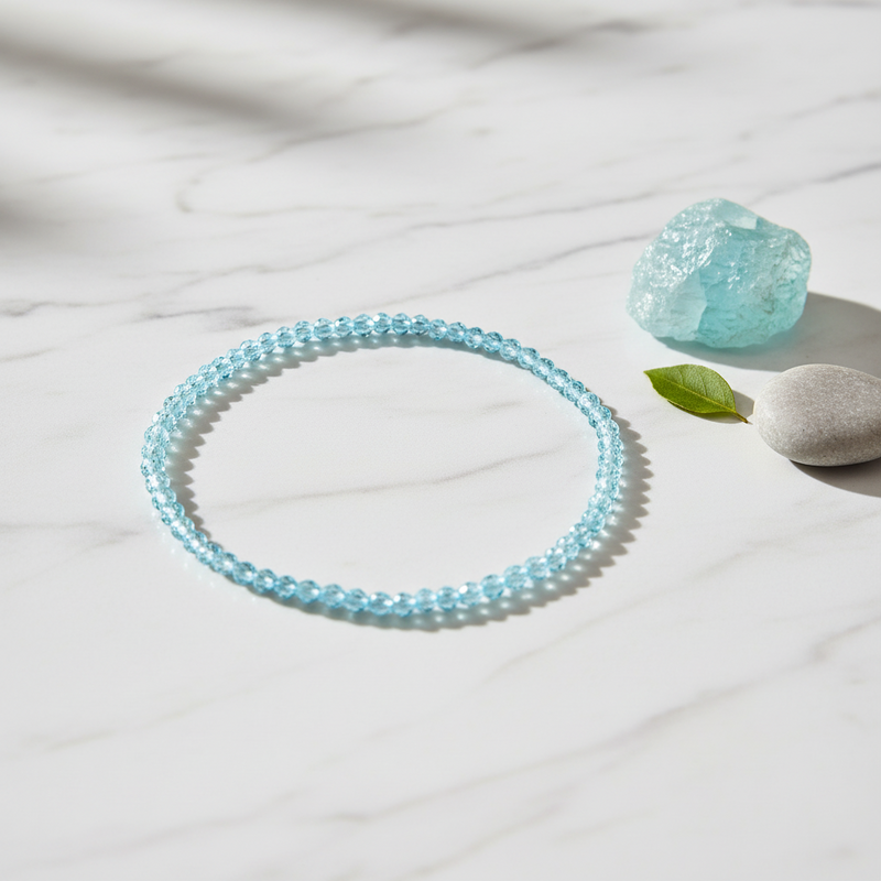 A light blue beaded bracelet arranged in a circle on a white marble surface, with a matching translucent blue stone and a smooth white stone nearby.