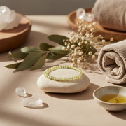 A light green beaded bracelet displayed on a smooth white stone, surrounded by spa-style props including rolled beige towels, dried flowers, green leaves, clear crystals, and a small bowl of oil on a warm beige surface.