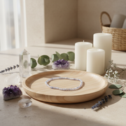 A beaded bracelet with pale lavender beads is displayed on a light wood tray, surrounded by white pillar candles, crystals, and green leaves in a warm, softly lit setting.