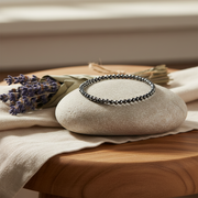 A beaded stretch bracelet with black beads, displayed on a round beige stone prop on a draped cream linen cloth over a wooden surface, with dried lavender sprigs nearby.