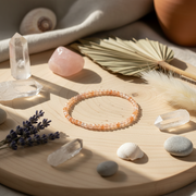 A beaded bracelet made of peach-orange translucent stones arranged in a circle on a light wood tray, surrounded by crystals, shells, dried lavender, a palm leaf, and soft neutral props in warm natural light.