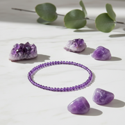 A purple beaded bracelet arranged in a circle on a white marble surface, surrounded by purple crystal stones and green eucalyptus leaves in soft natural light.