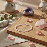 A delicate pink beaded bracelet rests on a light wooden board, styled with pink crystal clusters, rose petals, dried flowers, a smudge bundle, and a purple amethyst geode in a warm, sunlit setting.
