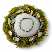 A beaded bracelet made of turquoise, blue, gray, and white beads, arranged in a circle on a smooth gray stone. The stone sits in the center of a circular arrangement of green moss, lavender sprigs, dried leaves, and clear crystals, all on a solid white background.