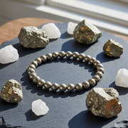 A beaded bracelet made of metallic silver-gray round beads is arranged in a circle on a dark slate tray, surrounded by rough white and metallic mineral stones in bright natural sunlight.