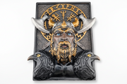 A Viking-themed decorative bookshelf decor featuring a sculpted horned warrior face with blue eyes, gold beard and helmet details, silver horns, two black ravens at the base, and a gold runic-style emblem on a dark textured rectangular panel, shown against a plain white background.