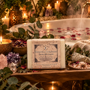 A bar of Spiritual Cleansing soap sits on a wooden table by a bath, surrounded by herbs, crystals and candles 