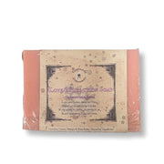 A packaged bar of soap wrapped in clear plastic, showing a tan paper label with decorative text and a pink soap bar visible on both sides, photographed on a white background.