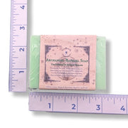 A rectangular bar of soap wrapped in clear plastic with a dusty pink paper label reading 'Archangels Raphael Soap,' shown on a white background with purple rulers measuring its width and height.