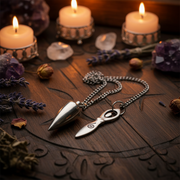 A silver-toned metal pendulum with a pointed conical weight and a small circular charm, displayed on a dark wooden surface with candles, crystals, dried lavender, and dried flowers.