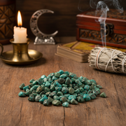 Emerald stones on a wooden surface with a candle, books, and smoldering sage in the background.