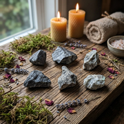 A styled arrangement of seven dark gray and black raw crystals on a rustic wooden table, surrounded by green moss, dried lavender sprigs, pink rose petals, a rolled burlap sack, and two lit beeswax candles.