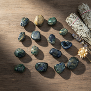 A set of polished dark blue and green tumbled stones arranged on a wooden surface, styled with a white sage bundle, dried lavender, and a small gold flower.