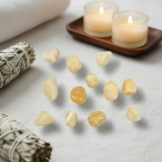 A set of small, polished yellow tumbled stones arranged on a white fabric surface, styled with a white sage bundle, a rolled white towel, and two lit tealight candles in the background.