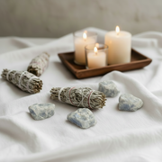 A styled arrangement of white sage smudge bundles tied with string, several smooth gray stones, and three lit white pillar candles in glass holders on a wooden tray, all set on draped white fabric.