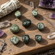 Greenish-brown stones on a wooden table in the sunshine with sage, and crystals around them.