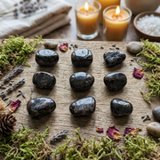 Nine polished black stones arranged in a 3x3 grid on a rustic wooden board, surrounded by green moss, dried lavender, small flowers, candles, and bath salts in a spa-like setting.