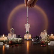 Hand holding a Rose Quartz Angel Pendulum over candles and crystals with a purple background