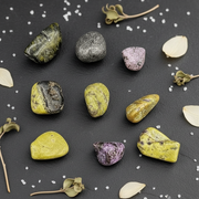 Nine polished tumbled stones in various colors and patterns are arranged on a dark slate background with scattered white sea salt and dried botanical elements.