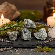 Three rough gray crystals arranged on a black slate tray with moss, rosemary, lavender sprigs, and lit candles in a cozy, blurred background.