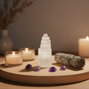 A white selenite tower crystal stands on a round wooden tray, surrounded by purple amethyst stones, a white sage smudge bundle, and three lit candles in glass holders against a softly blurred warm background.