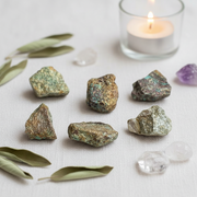 Seven raw, chrysocolla crystals with green, gold, and bronze hues are arranged on a textured white fabric surface, styled with dried olive leaves, clear quartz, a purple amethyst, and a lit white candle in a glass holder.