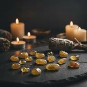 A group of polished yellow crystal stones arranged on a dark slate surface, with lit candles, a sage smudge bundle, and natural elements in the softly blurred background.