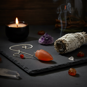 A carnelian pendulum , polished orange-red teardrop-shaped stone and a small red bead charm, displayed on a dark slate tray with a lit candle, white sage bundle, crystals, and smoky background elements.