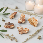 Five raw crystal formations with warm peach, orange, and brown hues are arranged on a white marble surface with decorative dried grasses, green leaves, and lit candles in the background.