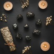 A styled flat-lay of polished black and white blizzard stone tumbled stones arranged on a dark slate surface, surrounded by a bundle of dried sage tied with twine, small white baby's breath flowers, and four lit tealight candles.