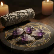 Four raw purple amethyst crystals arranged on a black circular tray with gold geometric line art, next to a white smudge bundle wrapped in string. Two lit candles are softly blurred in the background.