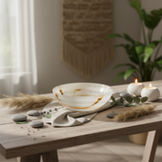 A white onyx bowl with brown marbled accents sits on a rustic wooden table styled with a beige cloth, green leaves, gray stones, dried grass, and two lit white candles in a softly lit interior setting.
