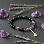 A beaded bracelet with glossy black round beads, silver-toned spacer beads, a cluster of small lavender-purple beads, and a broom-shaped charm, arranged on a dark gray surface with purple crystal points and lavender sprigs.