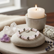 A beaded bracelet with pale pink round beads, silver spacer beads, and a small silver crescent moon charm, displayed on a smooth stone in a styled setting with a lit candle, crystals, and dried lavender.