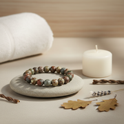 A beaded stretch bracelet made of marbled earthy beads in brown, green, and gray, displayed on a small round stone tray. The scene is styled with a white towel, a white candle, dried leaves, and twine on a neutral beige surface.