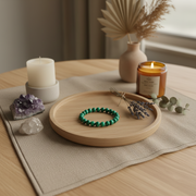 A green beaded bracelet is displayed on a round wooden tray, styled on a beige table runner with candles, crystals, and dried botanicals in a warm, cozy interior setting.
