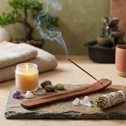 A wooden incense holder with a burning incense stick, styled on a stone tray with a lit candle, crystals, a smudge bundle, and folded towels in a serene tabletop setting.