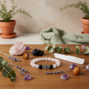 A beaded bracelet with pale pink, dark gray, and a central evil eye charm is displayed on a wooden table surrounded by crystals, eucalyptus leaves, lavender, and a white selenite wand.