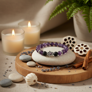 A beaded bracelet with purple, black, and white beads and a silver hamsa charm, displayed on a white stone atop a round wooden tray with candles, dried lotus pods, cinnamon sticks, and lavender.