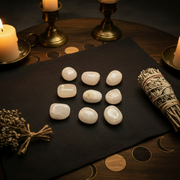 Nine polished pink calcite tumbled stones arranged on a black cloth, surrounded by lit candles, brass candlesticks, dried floral sprigs, and a bundle of white sage on a dark wooden surface.