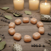 Ten smooth peach-colored stone cubes arranged in a circle on a rustic wooden surface, styled with lit candles, dried lavender sprigs, green leaves, and bundles of white sage.