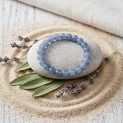 A light blue beaded bracelet displayed on a smooth gray stone, set in a styled arrangement with sand, sage leaves, lavender sprigs, and a folded cream fabric background.