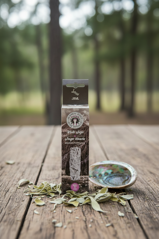 A boxed product labeled 'White Sage' stands upright on a rustic wooden surface outdoors, with scattered dried leaves in front and an abalone shell dish to the right. The background is a blurred forest scene.